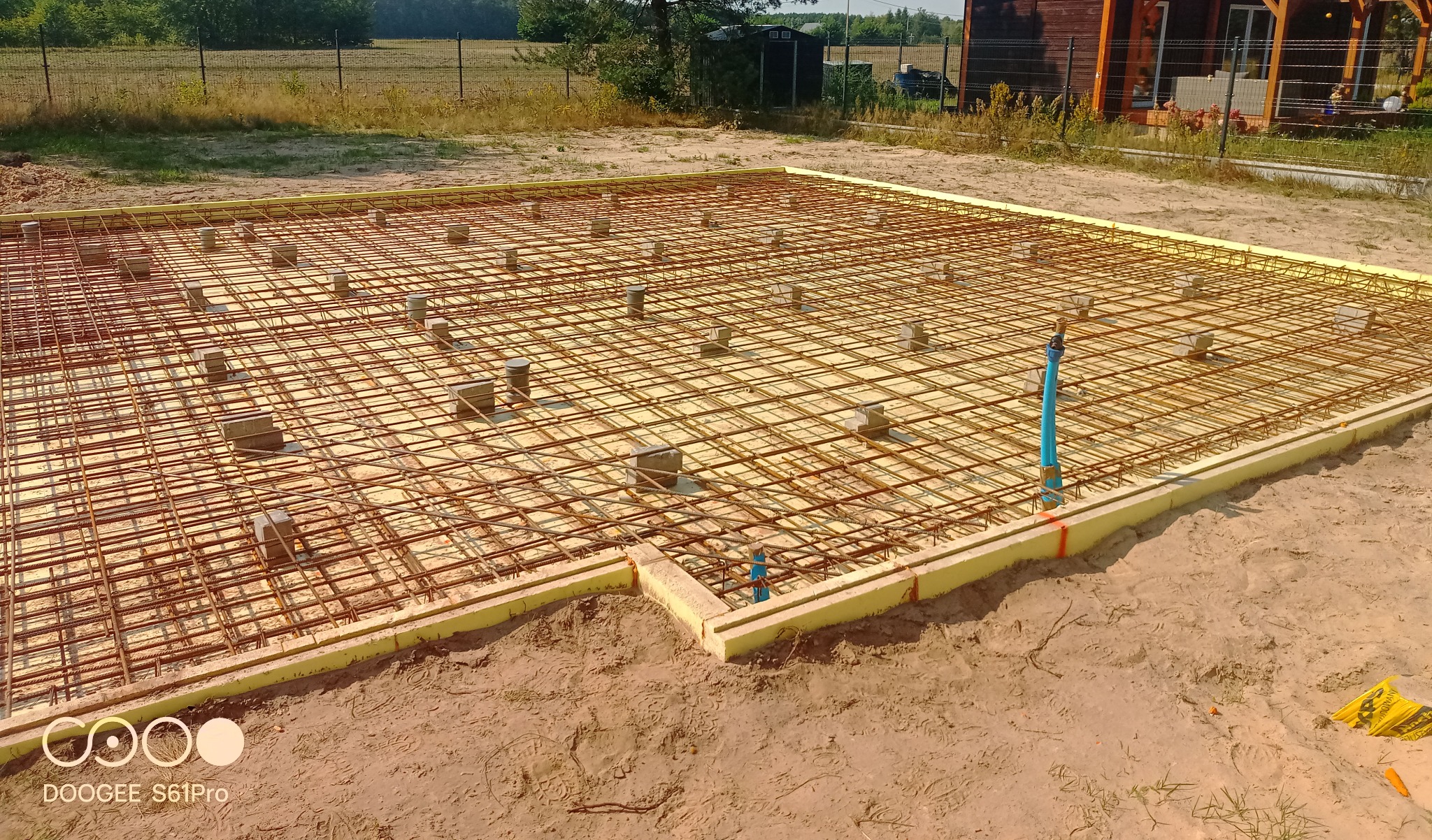 Zbrojenie fundamentu na piasku, z widocznymi podkładkami dystansowymi, szalunkiem z żółtego styropianu i wystającą rurą. W tle budynek i zieleń.