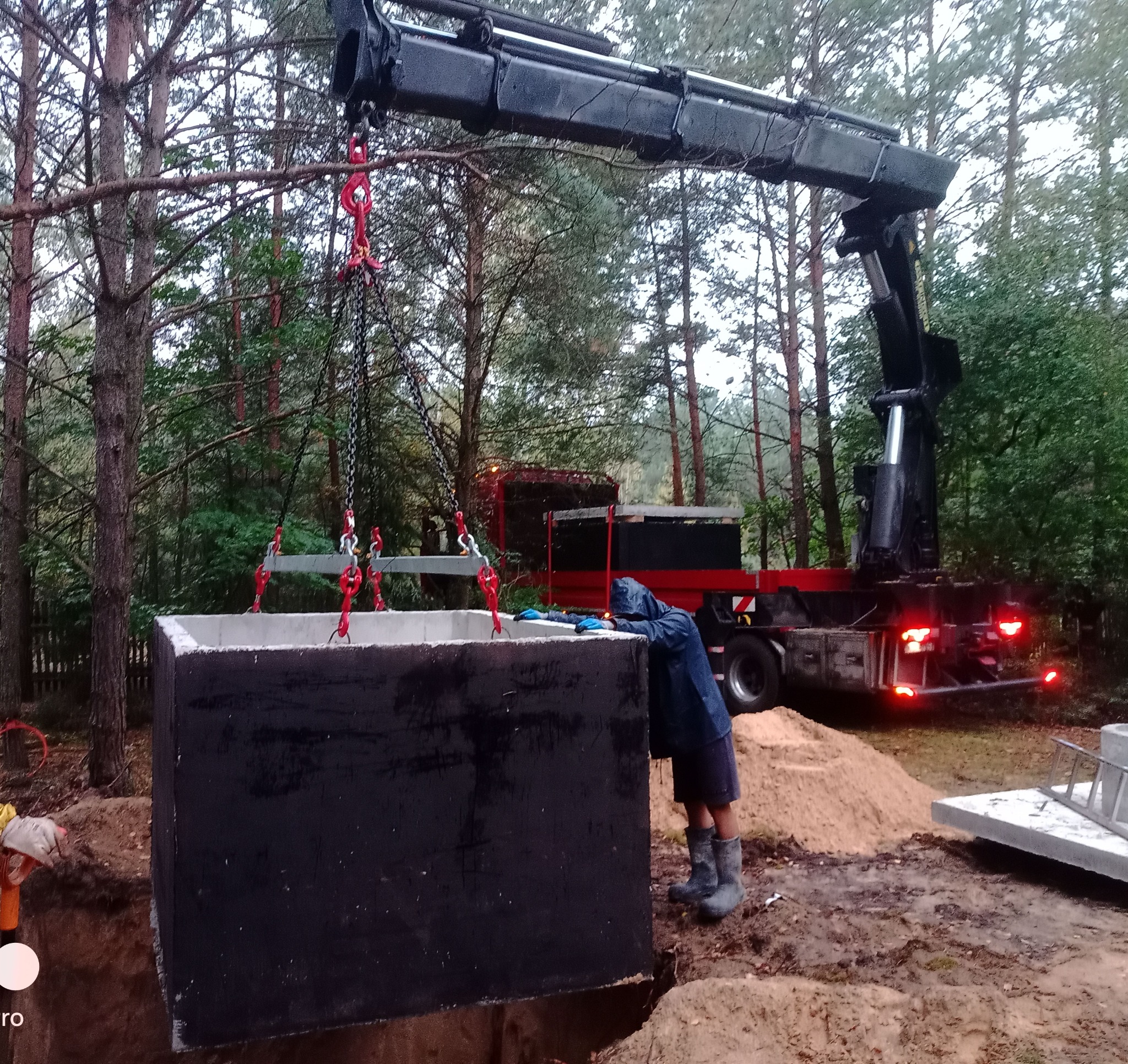 Montaż betonowego szamba za pomocą dźwigu na zalesionej działce. Pracownik w ubraniu roboczym nadzoruje opuszczanie zbiornika do wykopu. Widoczny samochód ciężarowy.