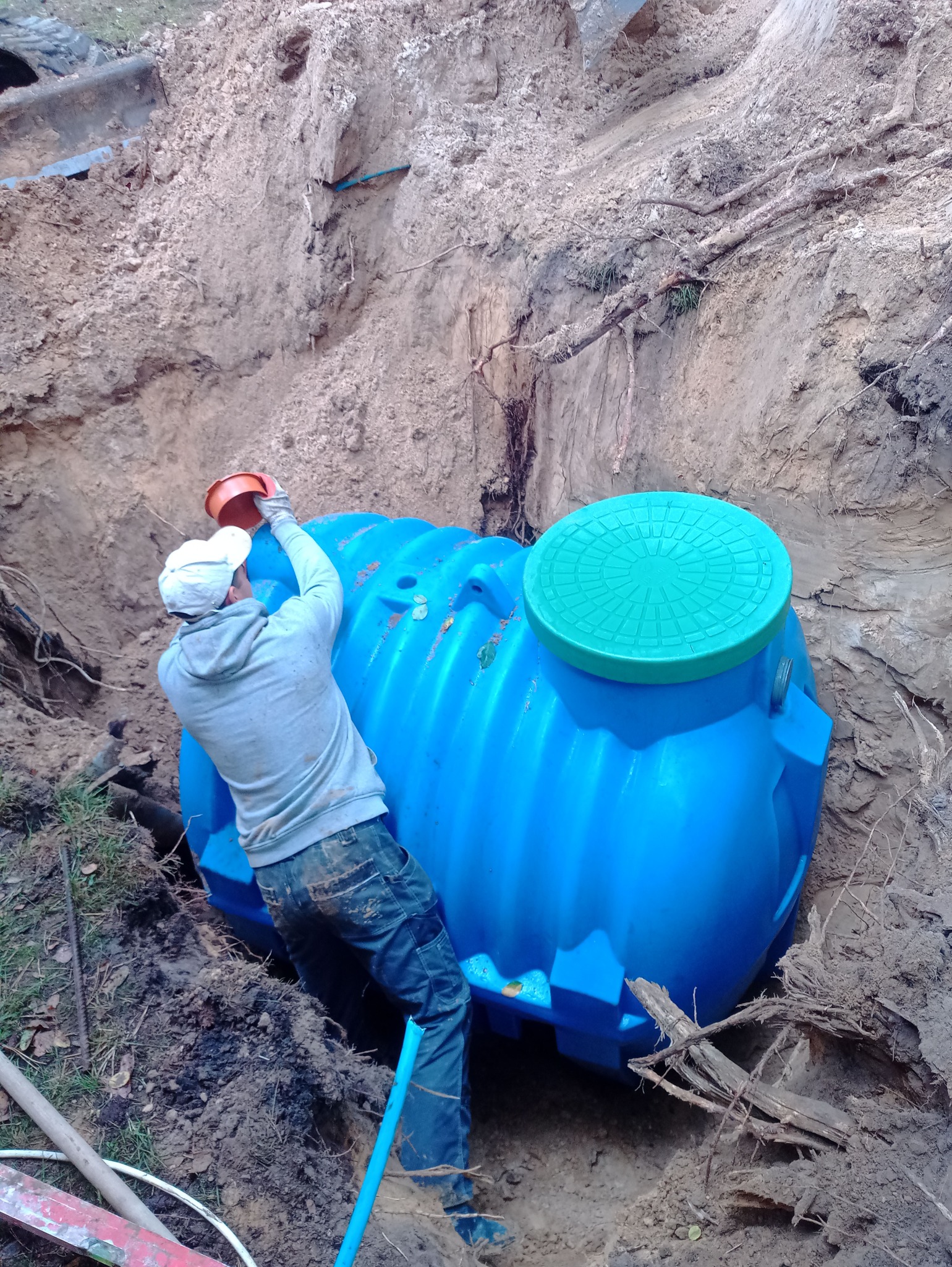Montaż niebieskiego zbiornika z zieloną pokrywą w wykopie. Pracownik w dresie i czapce zakłada pomarańczową pokrywę. Widoczne narzędzia i ziemia.