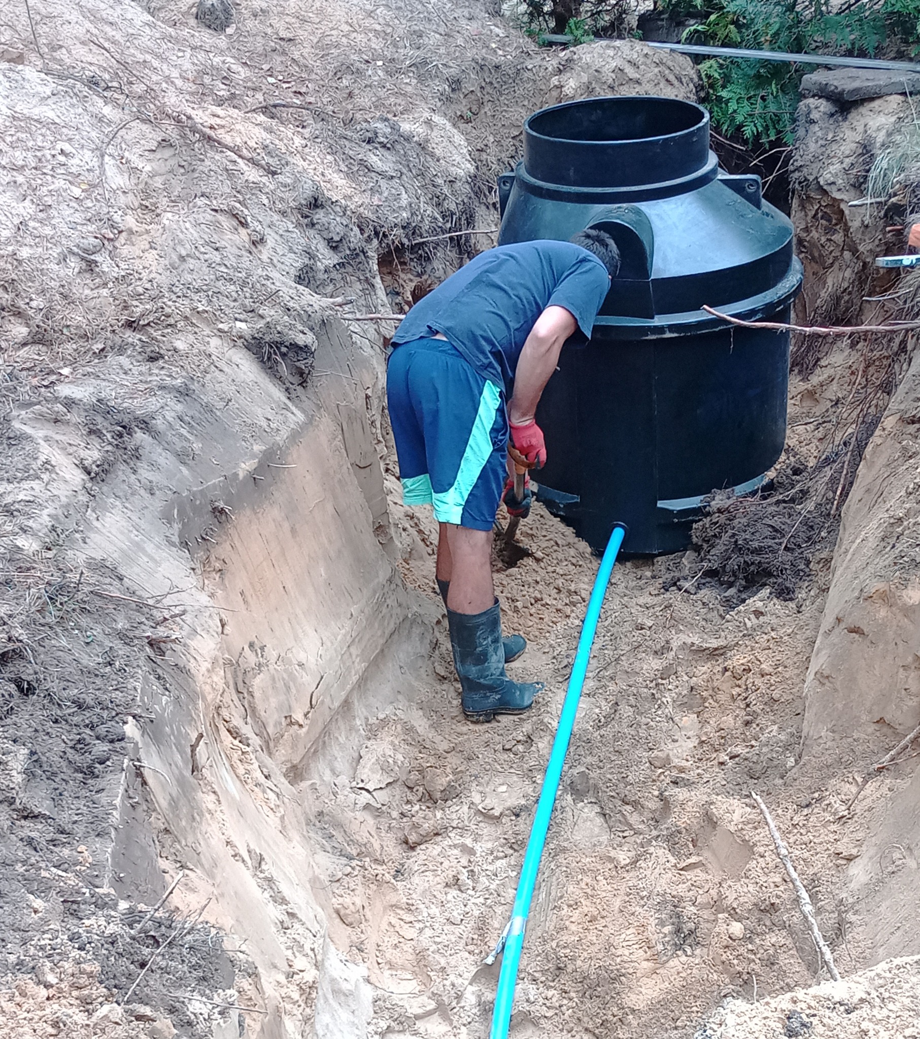 Montaż szamba: mężczyzna w wykopie, w gumowych butach, instaluje zbiornik z rurą. Prace ziemne w toku, widoczne ślady kopania, naturalne oświetlenie.