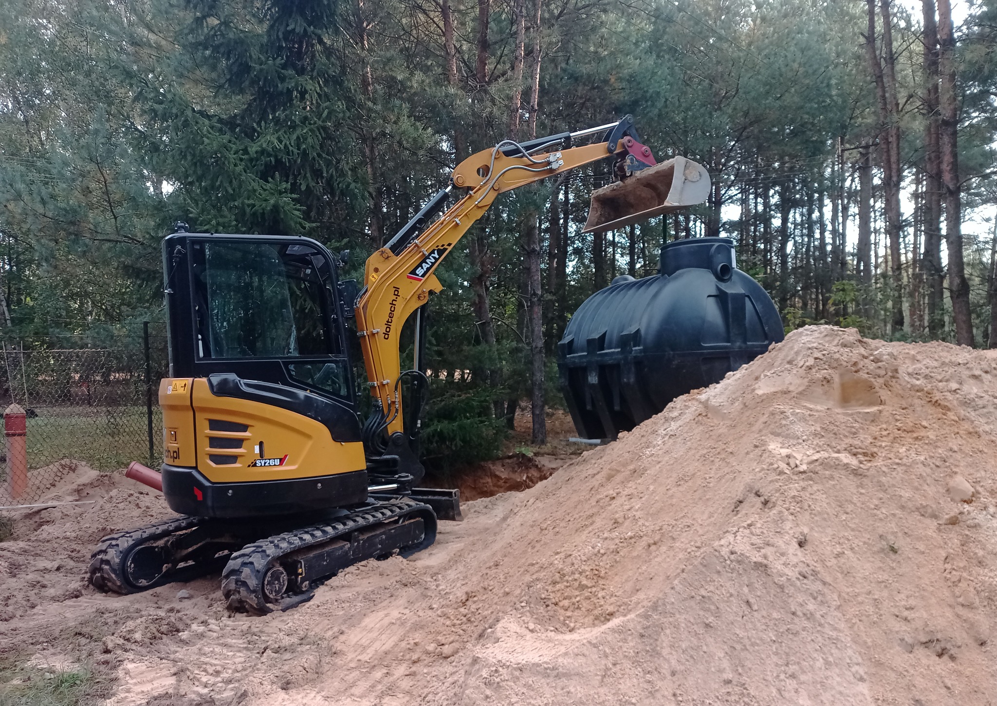 Żółta mini koparka SANY obok czarnego zbiornika na tle drzew. Wykopana ziemia tworzy spory nasyp. Widoczny etap instalacji zbiornika.