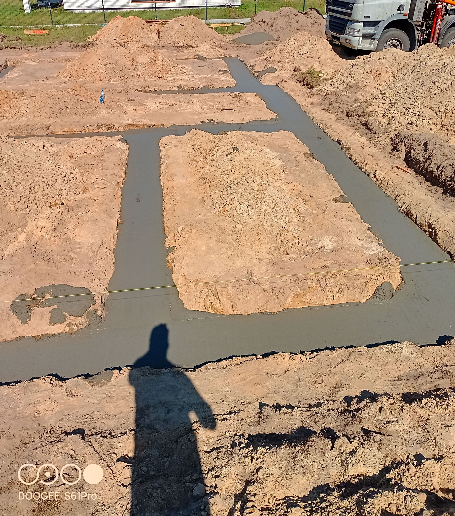 Wylane fundamenty pod budowę domu, widoczne wykopane rowy zalane betonem. W tle hałda ziemi i betoniarka. Cień osoby w rogu kadru.