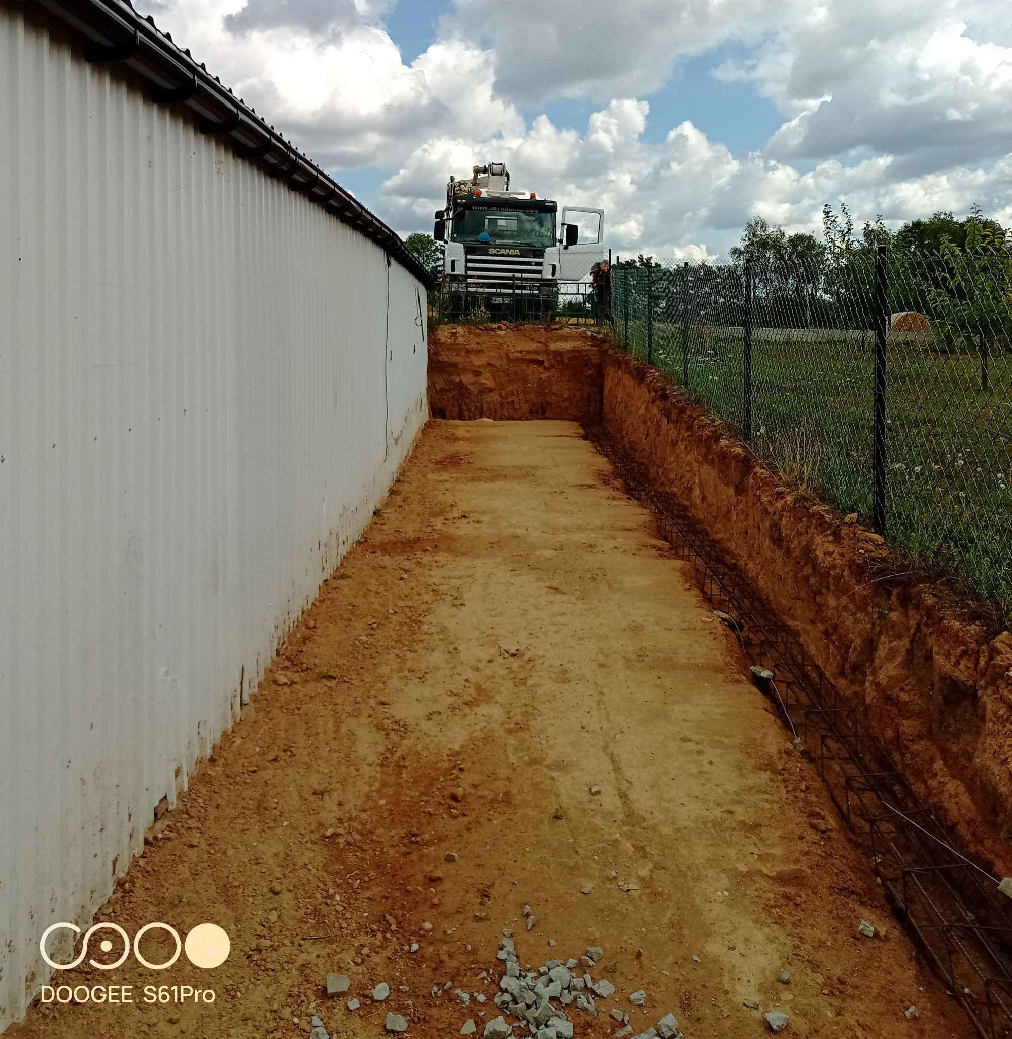 Wykop fundamentu z widocznym zbrojeniem i zaparkowaną koparką SCANIA w tle, na tle błękitnego nieba z chmurami. Widok z perspektywy gruntu.