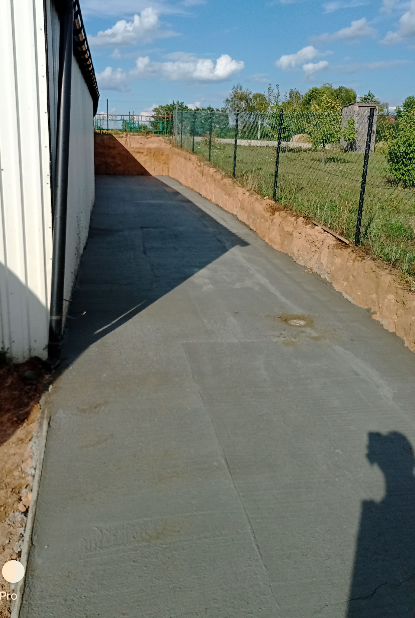 Świeżo wylana, gładka betonowa droga prowadząca wzdłuż budynku z blachy trapezowej, ogrodzona siatką. Widoczny wykopany grunt po bokach.