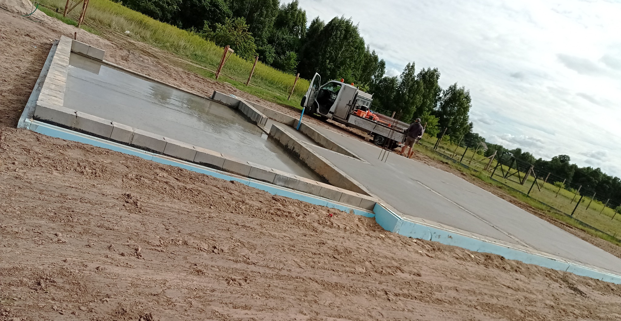 Wylane fundamenty z izolacją, obłożone bloczkami betonowymi, z widocznym pracownikiem i samochodem dostawczym na działce budowlanej w słoneczny dzień.