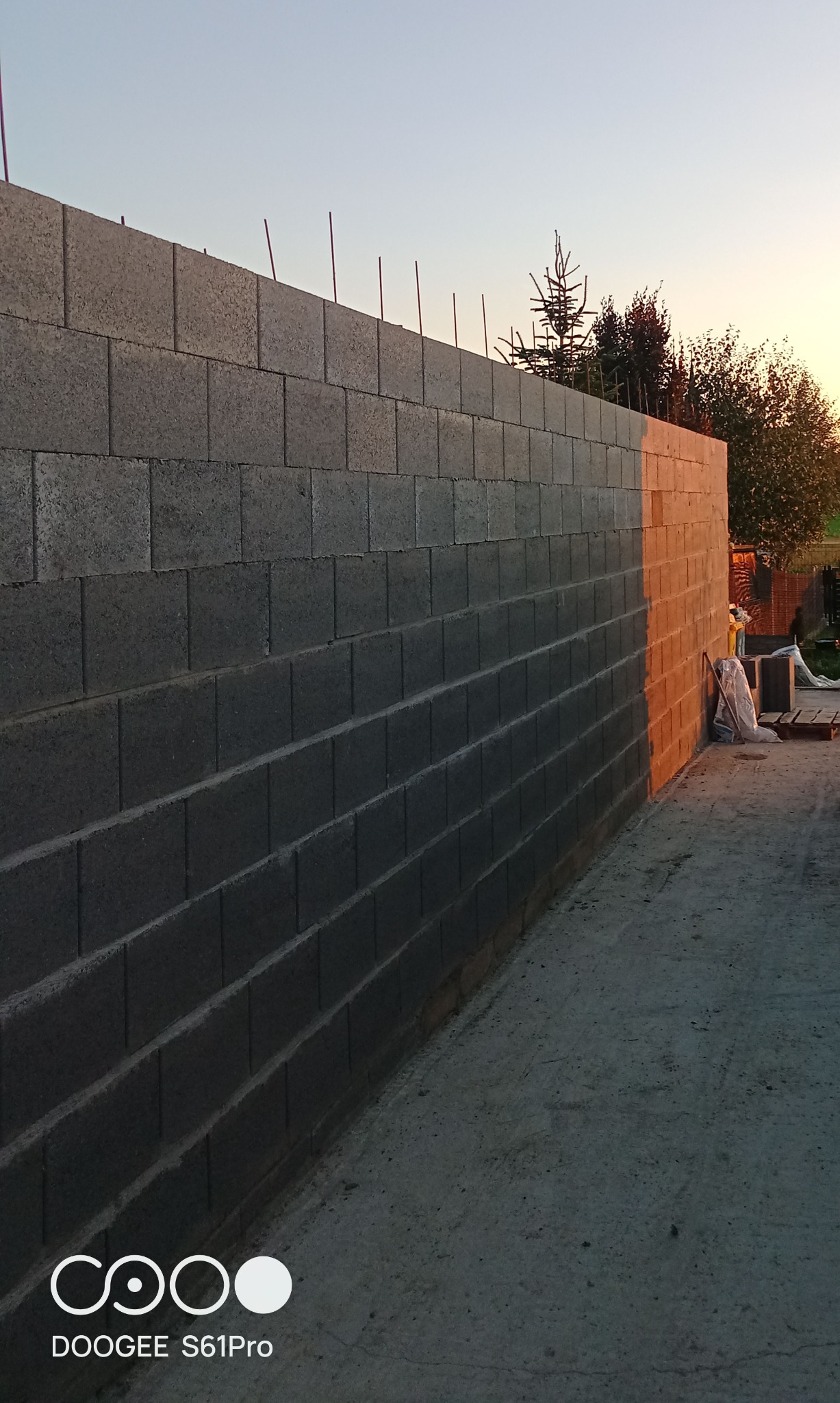 Ściana z szarego bloczka betonowego w trakcie budowy, wystające pręty zbrojeniowe, ciepłe światło zachodzącego słońca oświetla fragment ściany.