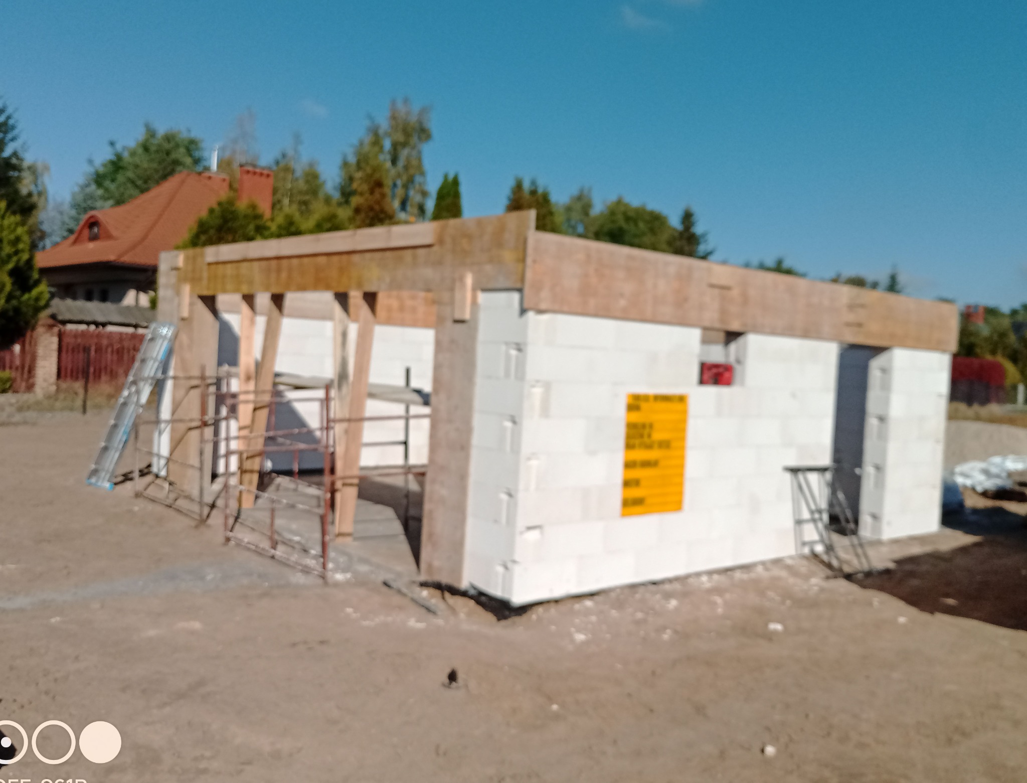 Budowa garażu z bloczków betonu komórkowego. Widoczne drewniane szalunki i rusztowania. W tle dom z czerwoną dachówką i zieleń drzew.
