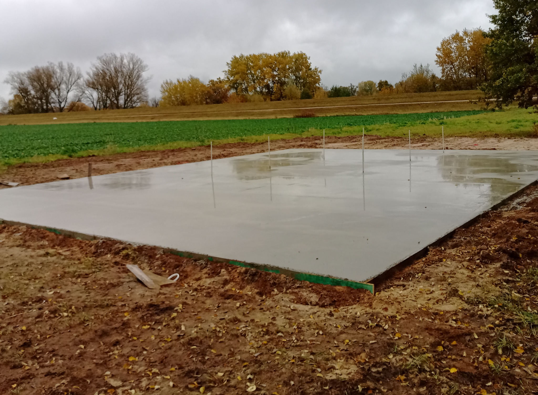 Świeżo wylany fundament pod budowę domu, widoczne szalunki i wystające pręty. W tle pole uprawne i drzewa pod pochmurnym niebem.