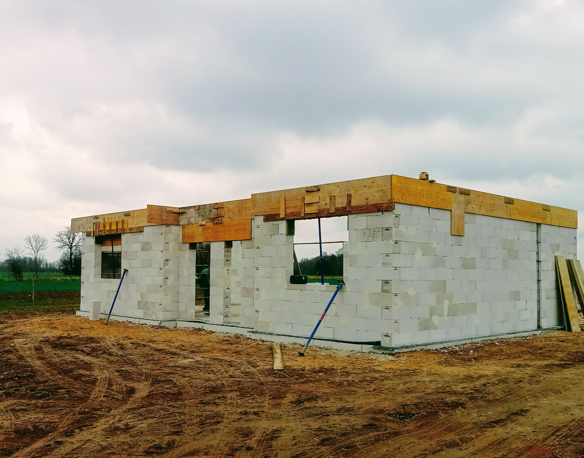Niedokończony budynek z białych bloczków, z widocznymi drewnianymi elementami konstrukcyjnymi stropu. Na pierwszym planie ziemia, w tle zieleń i pochmurne niebo.