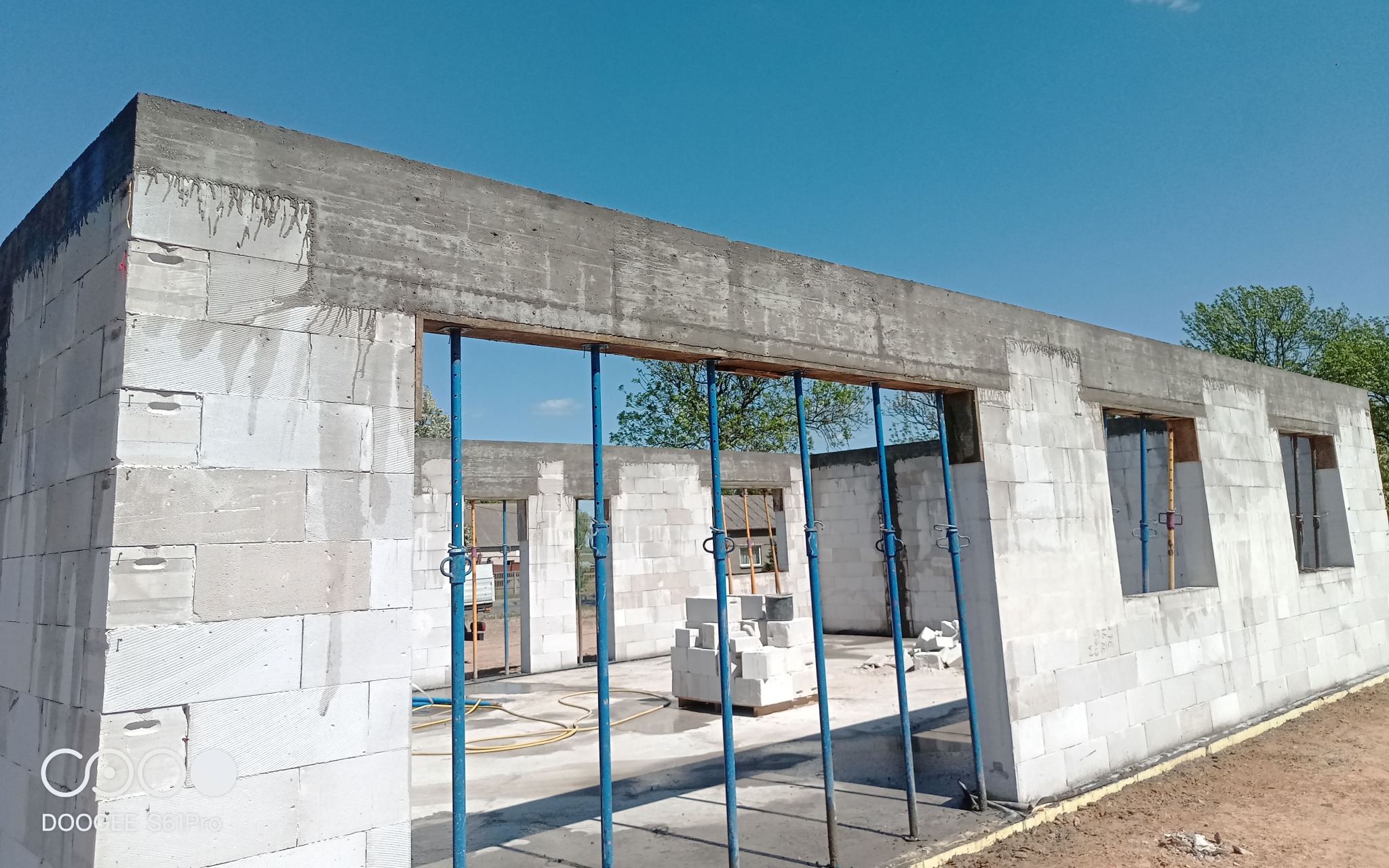 Budowa domu z betonu komórkowego: wzniesione ściany z otworami okiennymi, betonowy wieniec, podpory budowlane w kolorze niebieskim, błękitne niebo w tle.