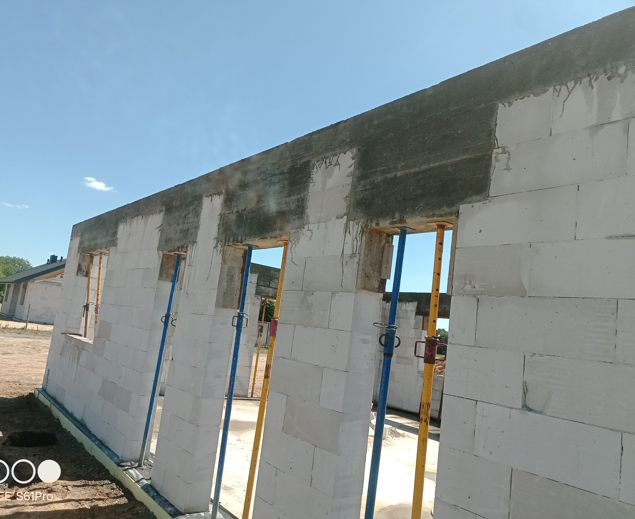 Ściana domu z betonu komórkowego w trakcie budowy, z widocznymi otworami okiennymi i betonowym wieńcem, podparta stemplami budowlanymi. Błękitne niebo w tle.