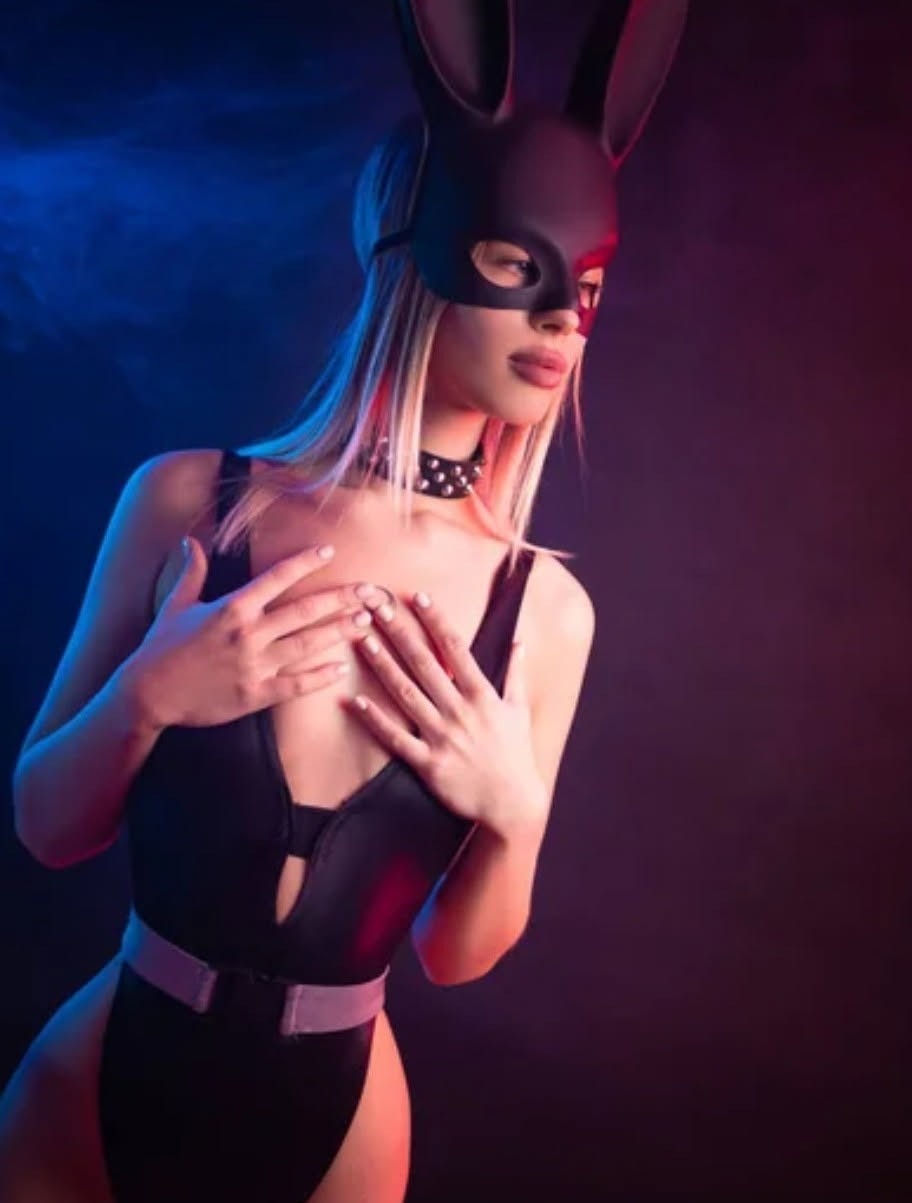 Kobieta w czarnym body i masce królika, oświetlona czerwono-niebieskim światłem, pozuje na ciemnym tle. Widoczny choker z ćwiekami i pasek.