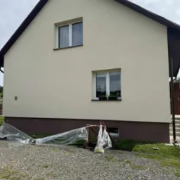 Dom z kremową elewacją i brązowymi akcentami. Widoczne okna z firankami i kwiatami. Na dole zabezpieczenia z folii malarskiej i rusztowanie.