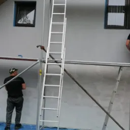 Dwóch mężczyzn na rusztowaniu maluje szarą elewację domu jednorodzinnego. Widoczna drabina i wiaderka z farbą. Częściowo pomalowana ściana.