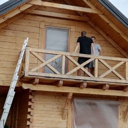 PatrON - Drewniany dom z balkonem w trakcie malowania. Widoczna drabina oparta o ścianę i dwie osoby na balkonie. Okna zabezpieczone folią. Czarny dach.