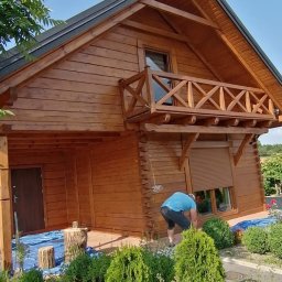 PatrON - Drewniany dom z balkonem, odświeżony nową warstwą lakieru, zabezpieczony folią malarską. Osoba przy oknie. Zieleń w ogrodzie. Słoneczny dzień.