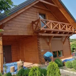 Drewniany dom z balkonem, odświeżony nową warstwą lakieru, zabezpieczony folią malarską. Osoba przy oknie. Zieleń w ogrodzie. Słoneczny dzień.