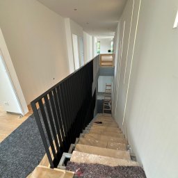 PatrON - Wnętrze domu w trakcie remontu: schody zabezpieczone, świeżo pomalowane ściany z wbudowanym oświetleniem liniowym. Czarna, metalowa barierka. Widoczna drabina.