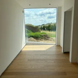 Nowoczesne wnętrze z dużym oknem, widok na krajobraz. Jasne ściany, drewniana podłoga. Minimalistyczny design, surowe wykończenie. Świeże, naturalne światło.