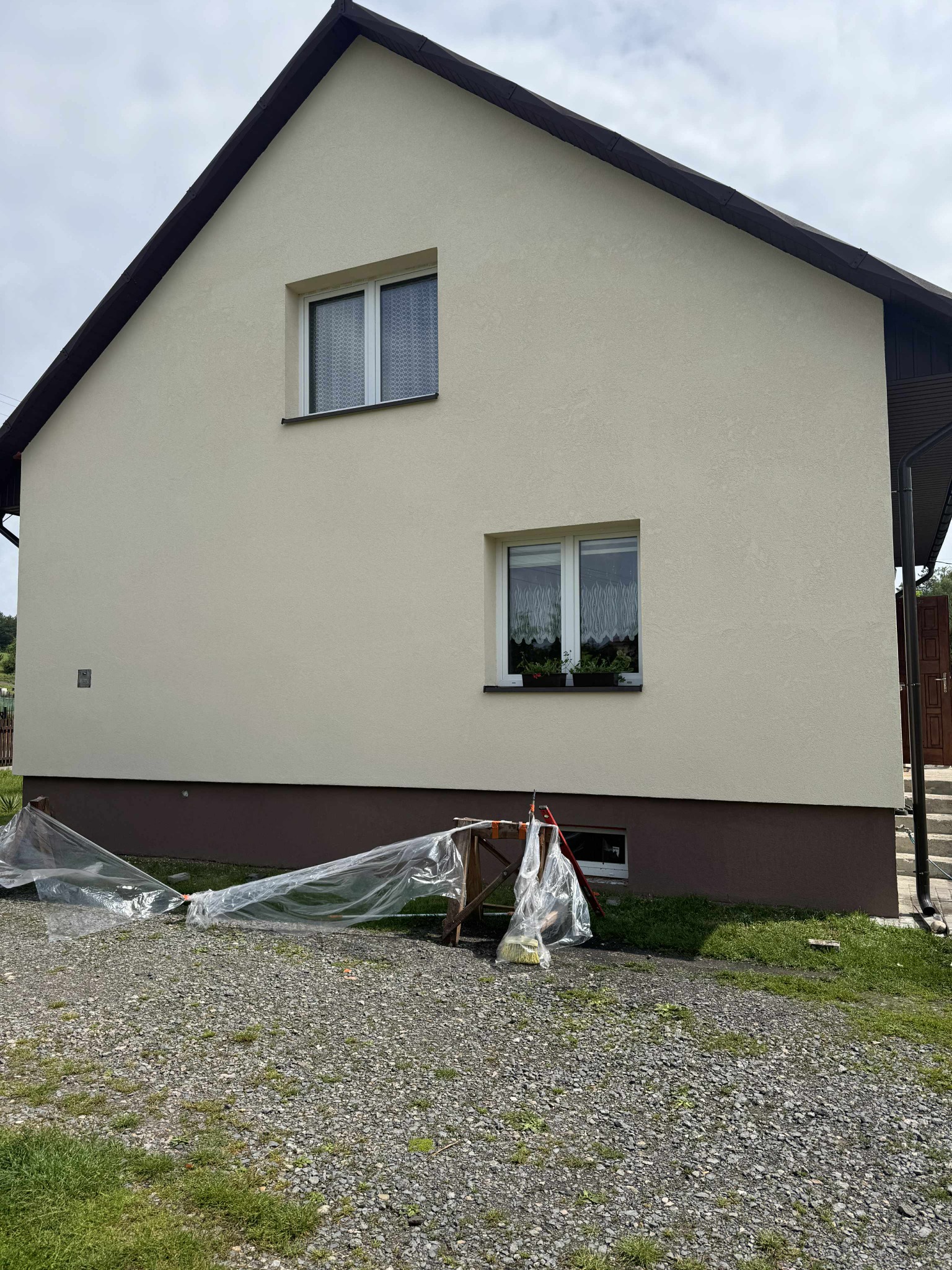 Dom z kremową elewacją i brązowymi akcentami. Widoczne okna z firankami i kwiatami. Na dole zabezpieczenia z folii malarskiej i rusztowanie.