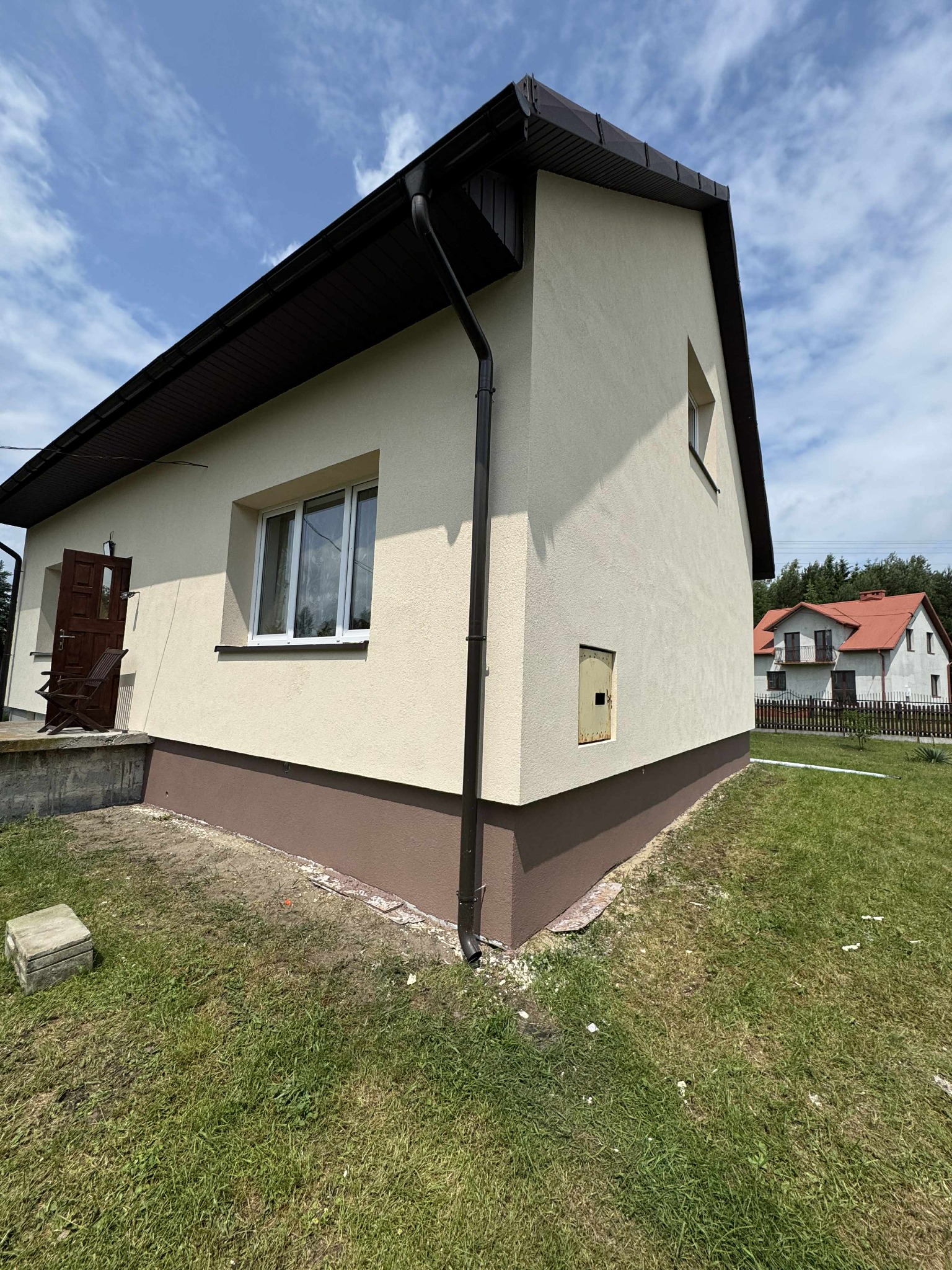 Wykończony budynek jednorodzinny z beżową elewacją i brązowym cokołem, widok z boku na tle błękitnego nieba. Wyraźne detale wykończenia, trawnik.