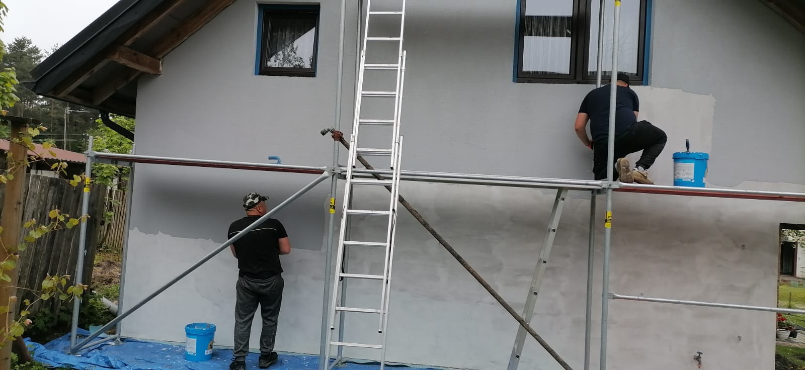 Dwóch mężczyzn na rusztowaniu maluje szarą elewację domu jednorodzinnego. Widoczna drabina i wiaderka z farbą. Częściowo pomalowana ściana.
