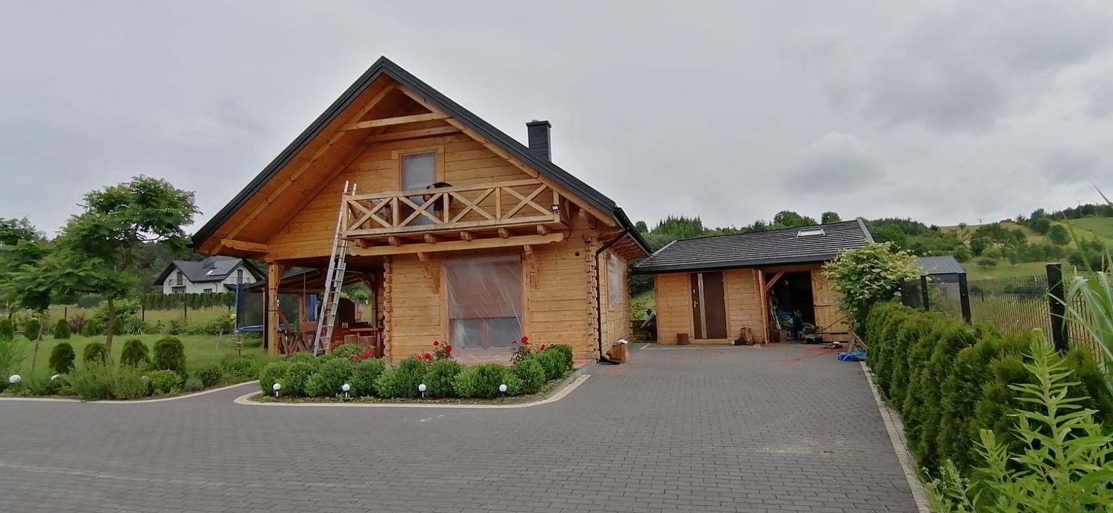 Drewniany dom z poddaszem i balkonem, częściowo osłoniętym folią. Z tyłu widoczny budynek gospodarczy, a obok drabina oparta o ścianę. Całość otoczona zielenią i brukowanym podjazdem.