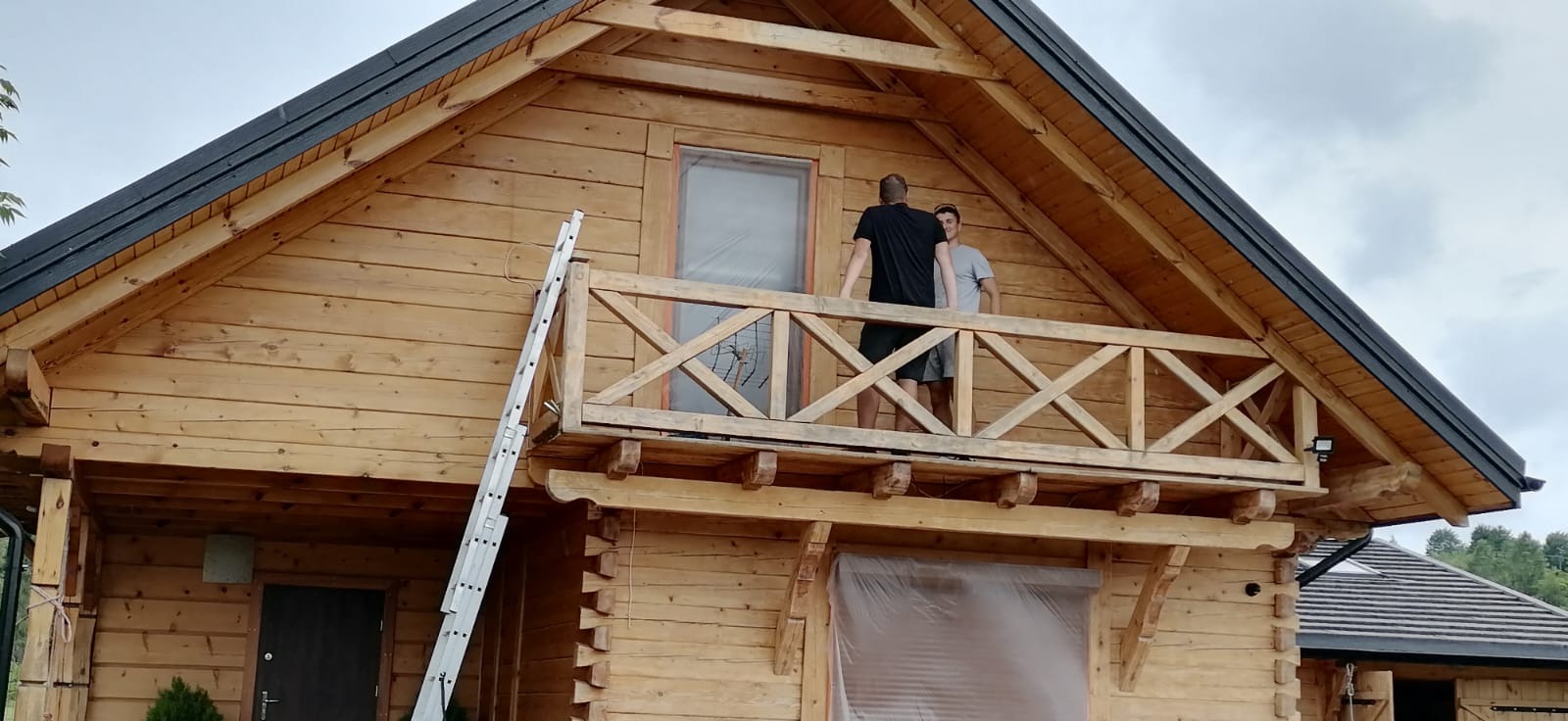 Drewniany dom z balkonem w trakcie malowania. Widoczna drabina oparta o ścianę i dwie osoby na balkonie. Okna zabezpieczone folią. Czarny dach.