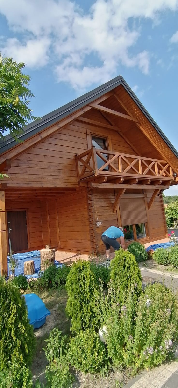 Drewniany dom z balkonem, odświeżony nową warstwą lakieru, zabezpieczony folią malarską. Osoba przy oknie. Zieleń w ogrodzie. Słoneczny dzień.