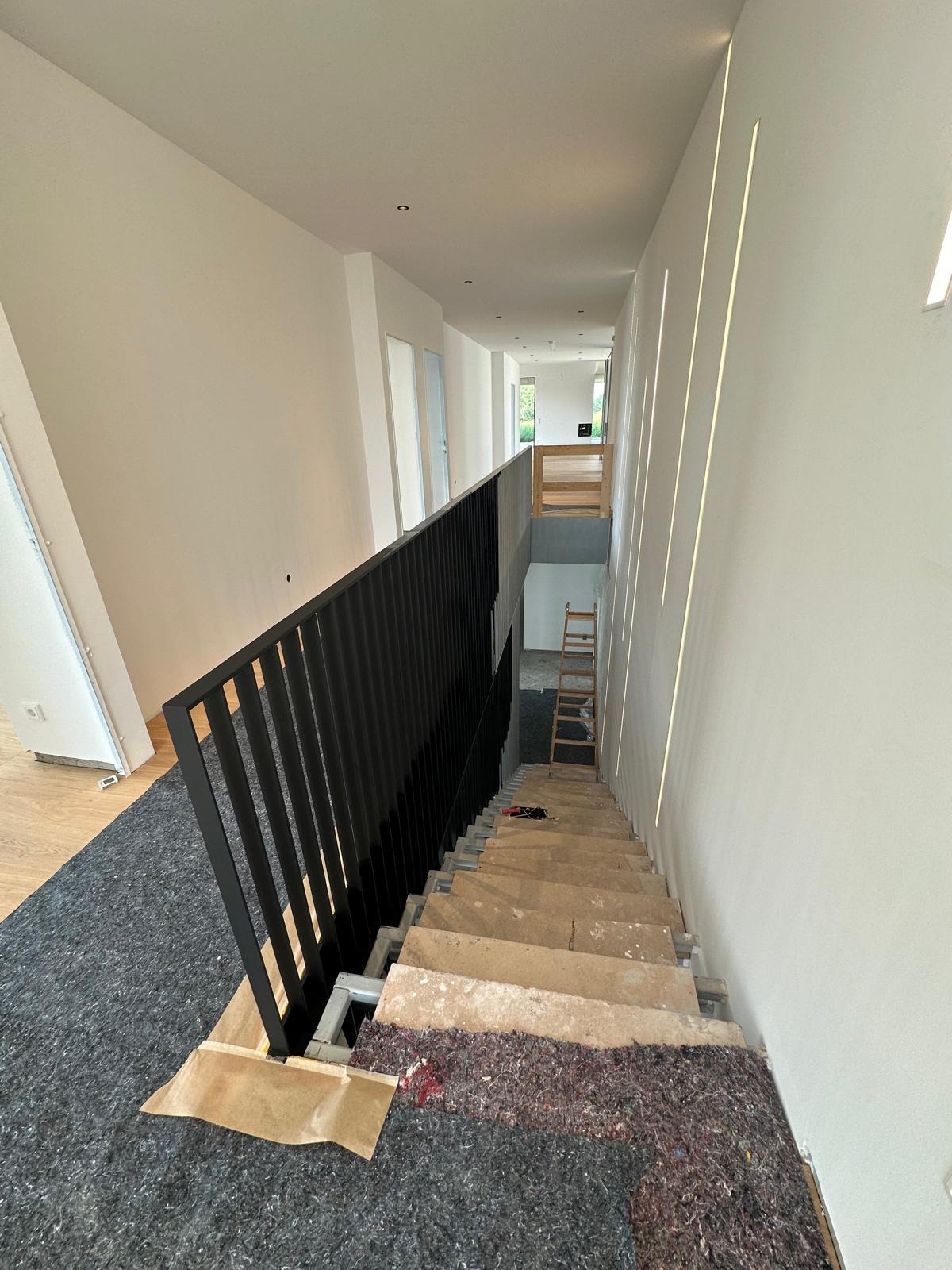 Wnętrze domu w trakcie remontu: schody zabezpieczone, świeżo pomalowane ściany z wbudowanym oświetleniem liniowym. Czarna, metalowa barierka. Widoczna drabina.