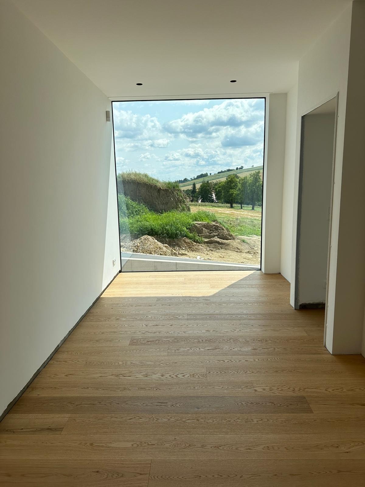 Nowoczesne wnętrze z dużym oknem, widok na krajobraz. Jasne ściany, drewniana podłoga. Minimalistyczny design, surowe wykończenie. Świeże, naturalne światło.