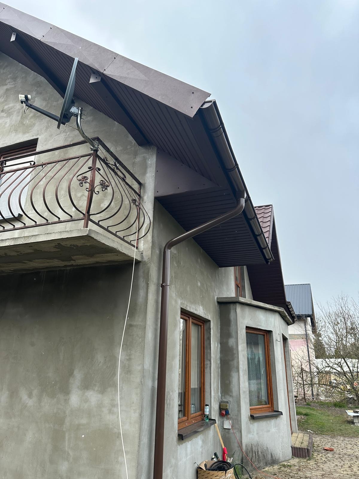 Surowa elewacja domu z kutym balkonem i brązowymi rynnami, widok z dołu na szarą ścianę i brązowe elementy wykończeniowe. Satelita na balkonie.