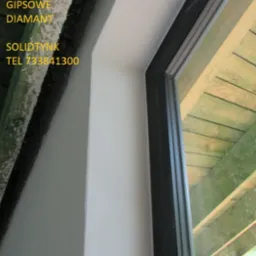 Gładko wykończone wnętrze okna z nową ramą. Widoczne świeżo położone, białe wykończenie. Fragment drewnianej konstrukcji dachu w tle.