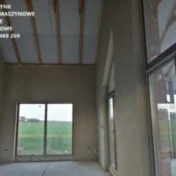 Surowe, jasne wnętrze domu po nałożeniu tynków. Widoczne okna z widokiem na zewnątrz. Na suficie widoczne drewniane elementy konstrukcyjne.