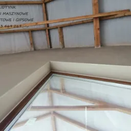 Świeżo otynkowane poddasze z widoczną konstrukcją więźby dachowej i oknem dachowym. Na ścianie widnieje napis reklamowy firmy tynkarskiej.