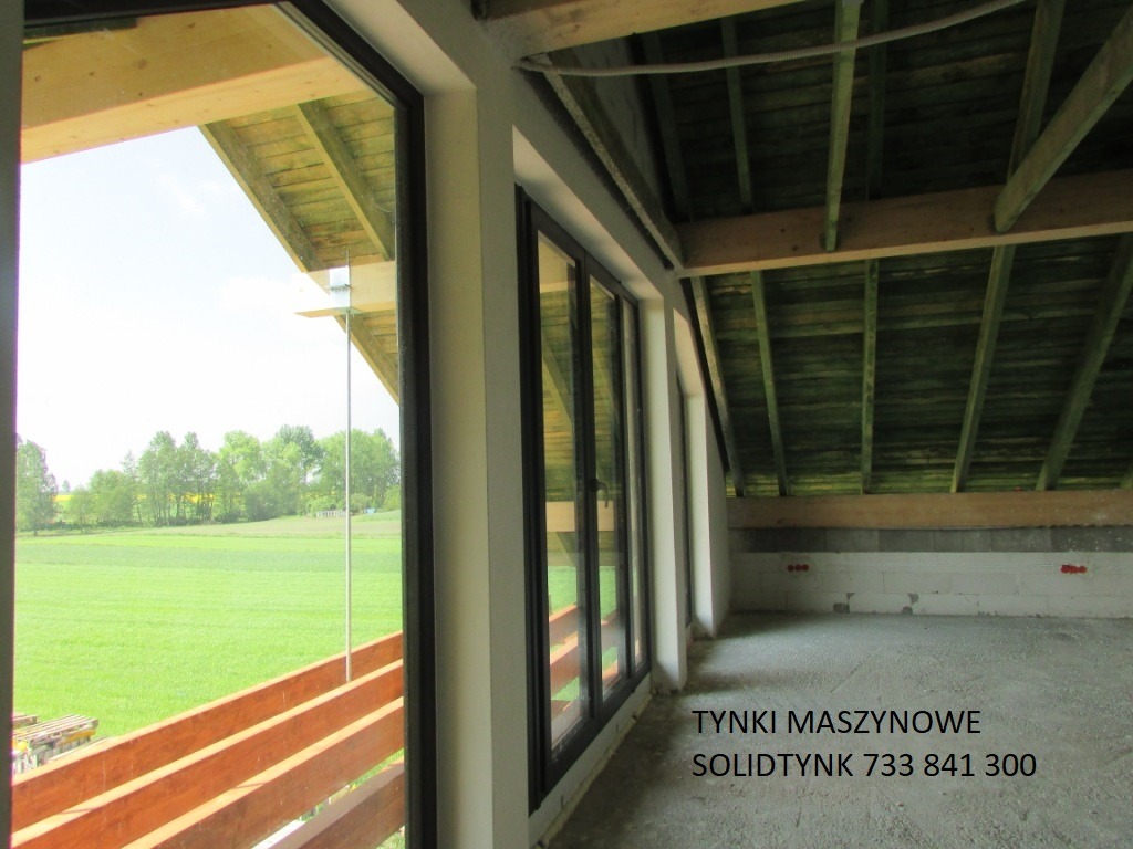 Surowy stan poddasza z widokiem na zewnątrz przez duże okna. Widoczne elementy konstrukcji dachu i ściany z pustaków. Jasne wnętrze z surowym wykończeniem.