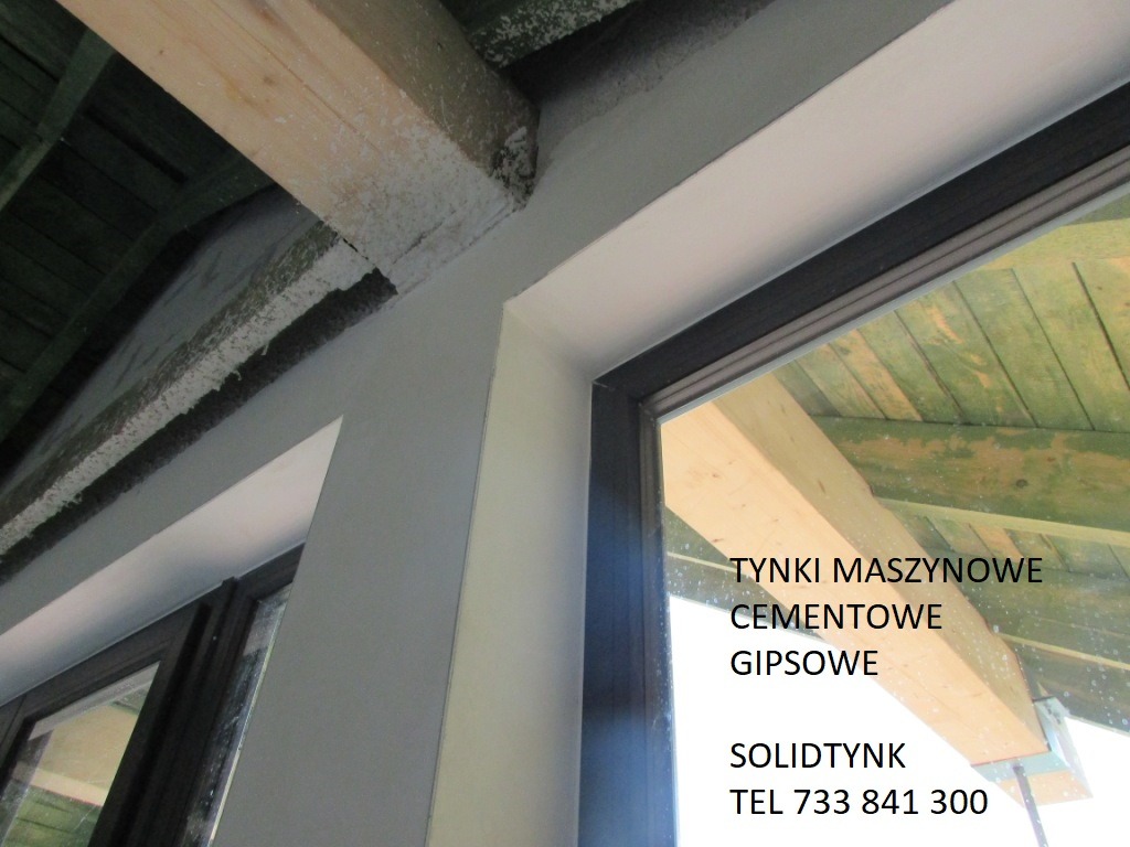 Szare, świeżo położone tynki przy oknie z widoczną drewnianą konstrukcją dachu. Reklama firmy z nazwą i numerem telefonu widoczna w odbiciu szyby.