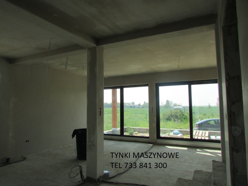 Surowe wnętrze domu w stanie deweloperskim: ściany po tynkowaniu maszynowym, duże okna z widokiem na zieleń, betonowa podłoga. Widoczny worek i kable na podłodze.