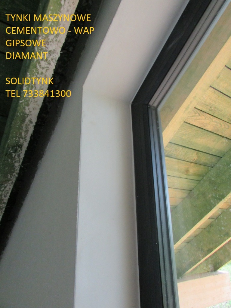 Gładko wykończone wnętrze okna z nową ramą. Widoczne świeżo położone, białe wykończenie. Fragment drewnianej konstrukcji dachu w tle.