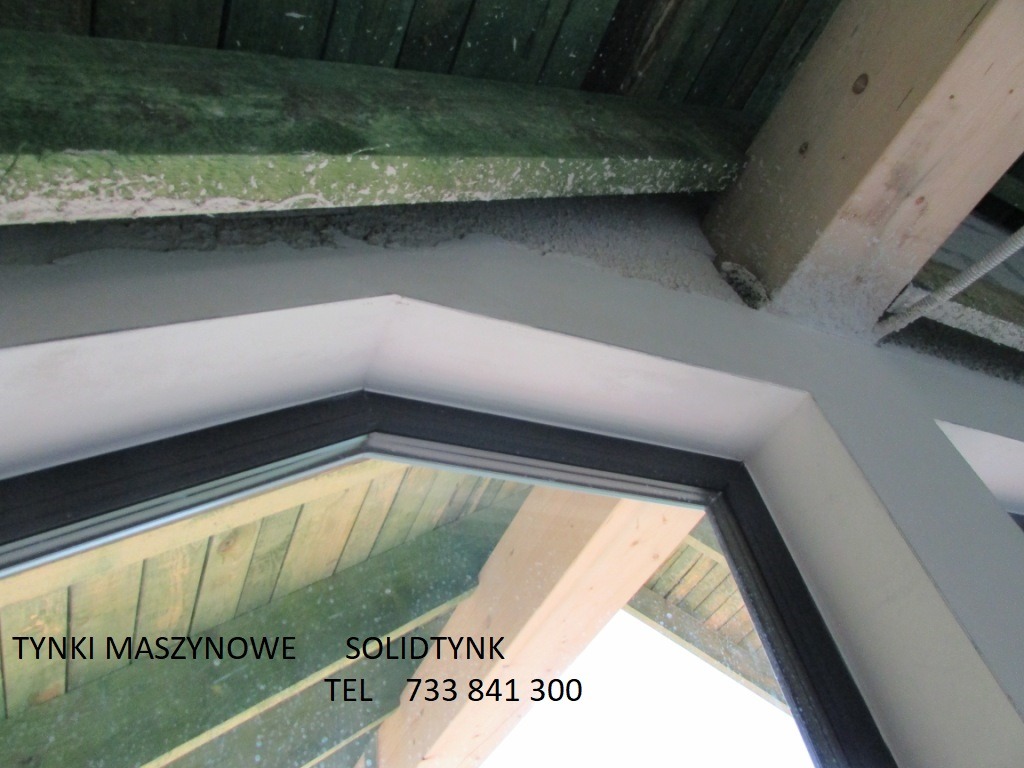 Wykończenie okna tynkiem w surowym wnętrzu. Widoczne drewniane elementy konstrukcyjne i biała rama okienna z czarnym obramowaniem. Świeżo położony tynk na ścianie.