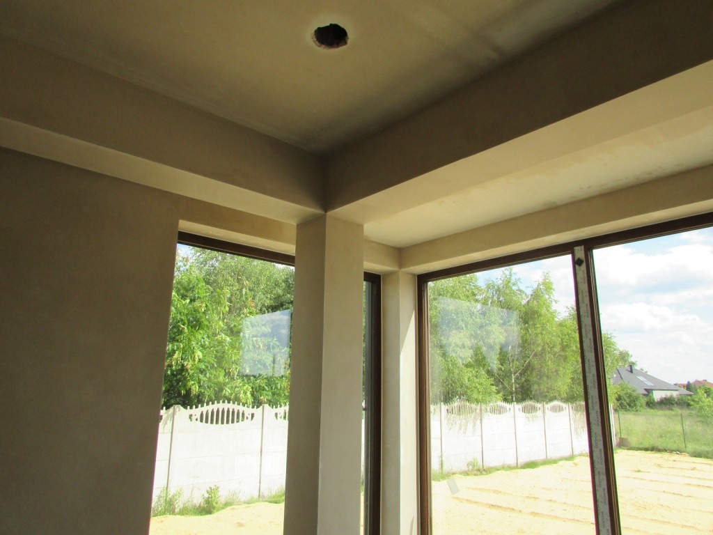 Wnętrze z oknami: gładkie, beżowe ściany i sufit po tynkowaniu, widoczne łączenia. Na zewnątrz zieleń drzew i białe ogrodzenie. Otwór na lampę w suficie.