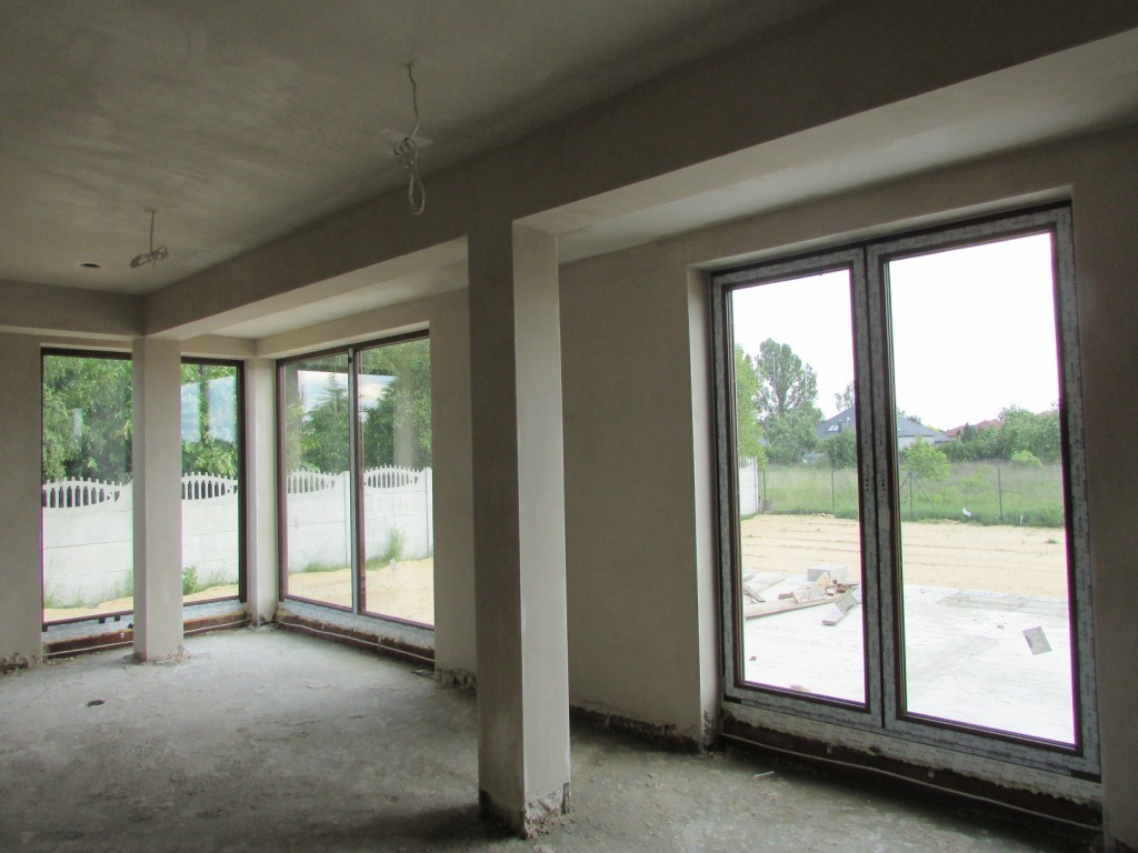 Surowy stan wnętrza z dużymi oknami i kolumnami. Widoczne tynki na ścianach i suficie, przygotowane do dalszych prac wykończeniowych. Stan deweloperski.