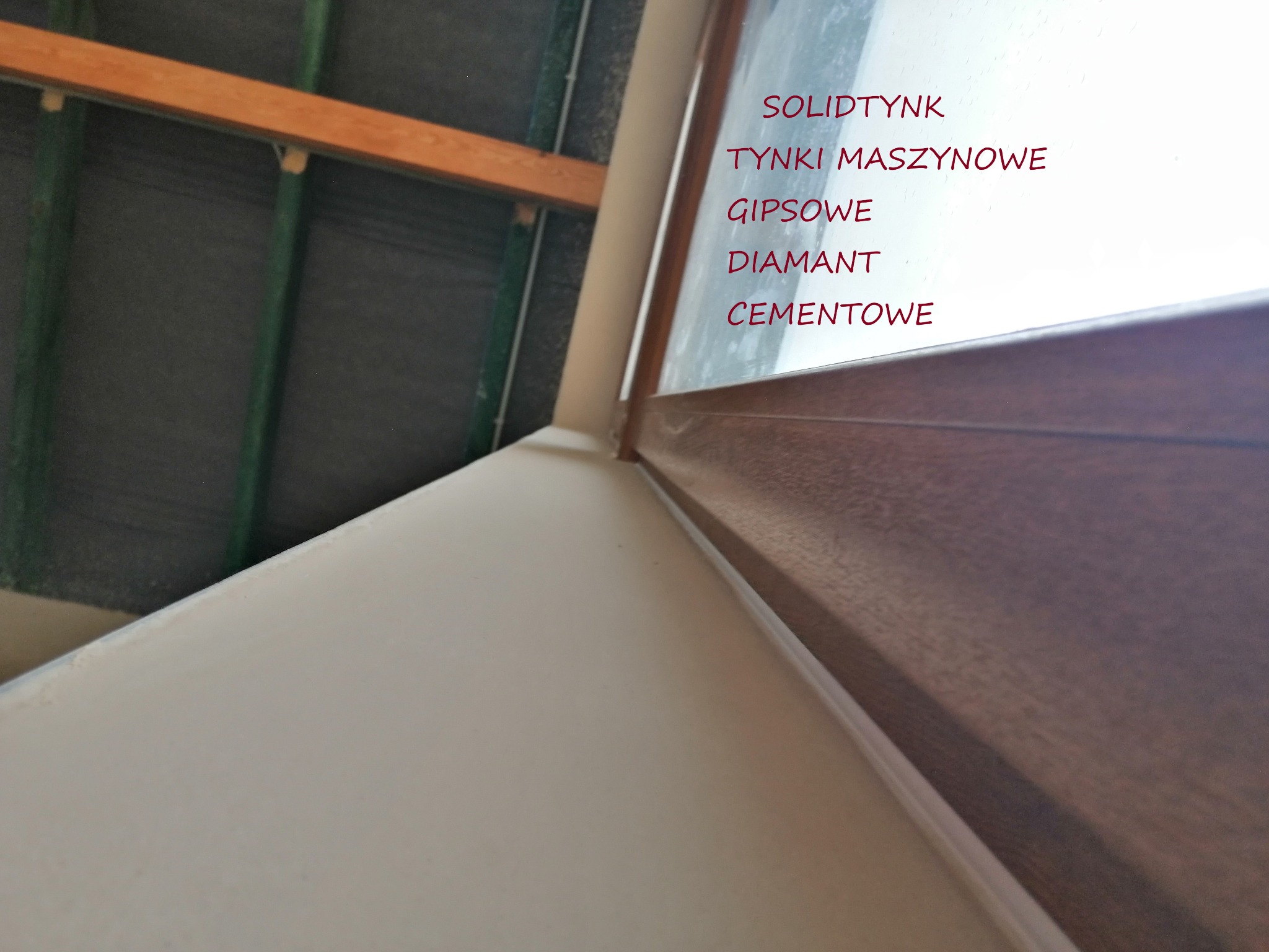 Świeżo położony, jasny tynk na ścianie przy oknie z drewnianą ramą, widoczne elementy konstrukcji dachu. Tekst reklamowy na szybie okna.