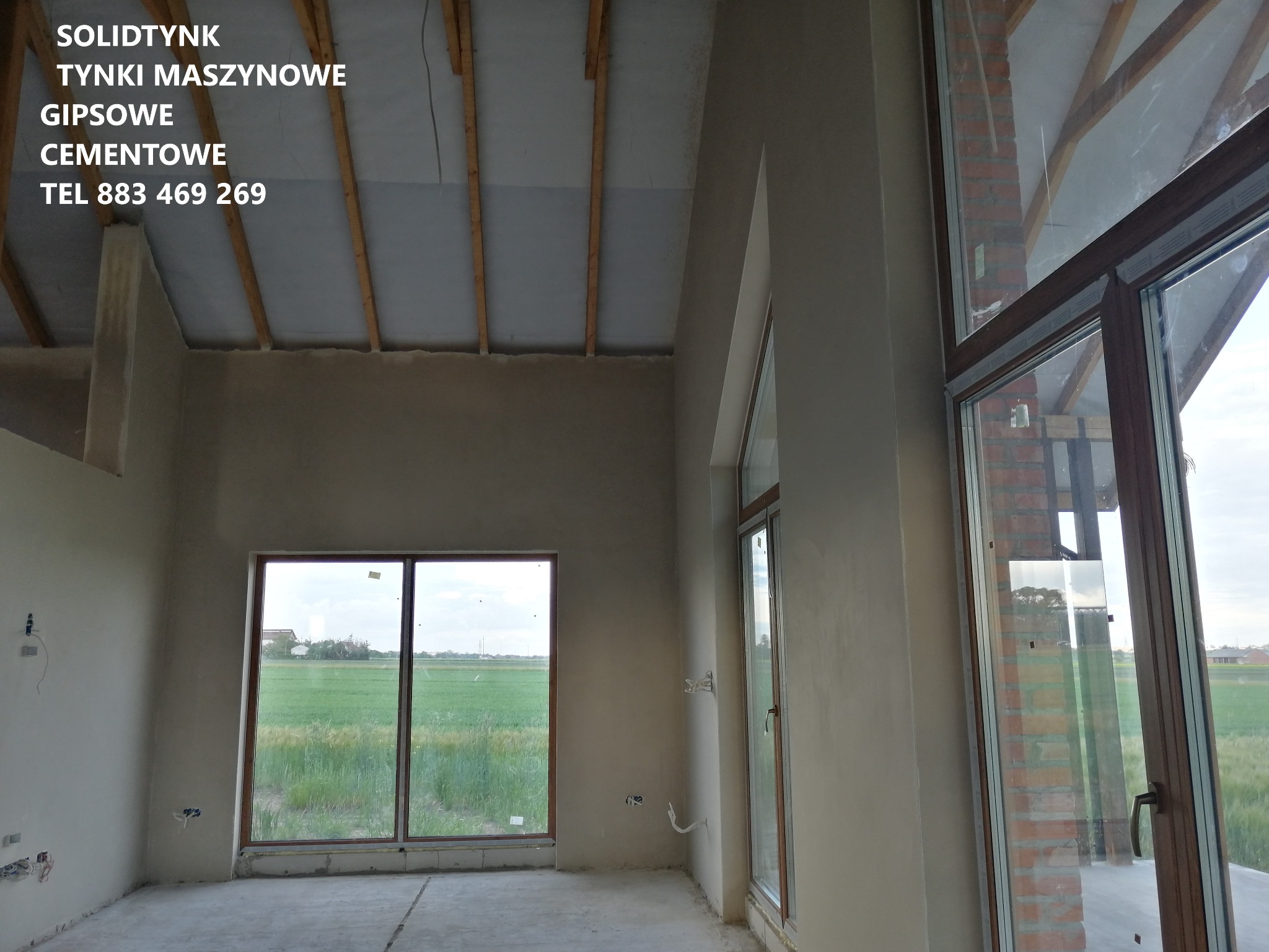 Surowe, jasne wnętrze domu po nałożeniu tynków. Widoczne okna z widokiem na zewnątrz. Na suficie widoczne drewniane elementy konstrukcyjne.