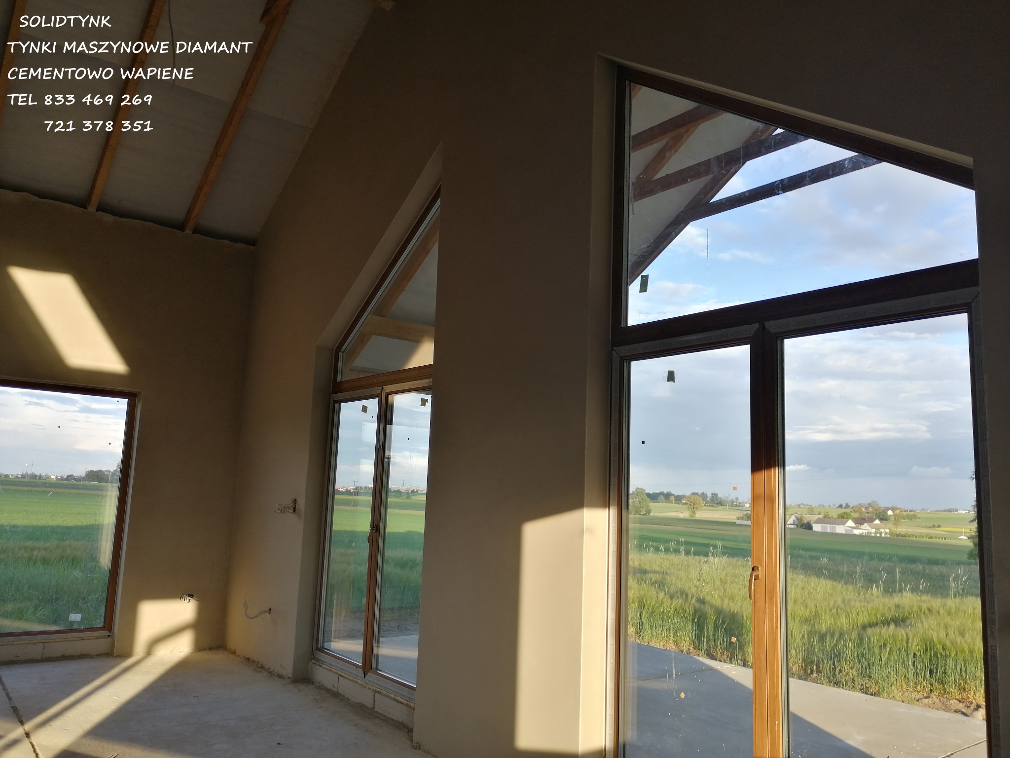 Wnętrze domu w trakcie prac wykończeniowych: świeżo położone jasne tynki na ścianach, duże okna z widokiem na zielone pola. Widoczne niezabezpieczone kable elektryczne.