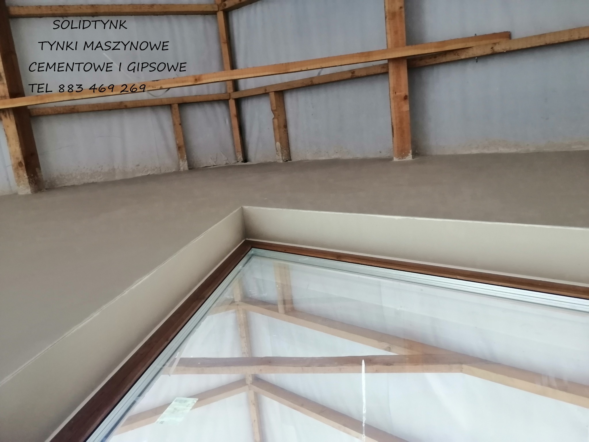 Świeżo otynkowane poddasze z widoczną konstrukcją więźby dachowej i oknem dachowym. Na ścianie widnieje napis reklamowy firmy tynkarskiej.