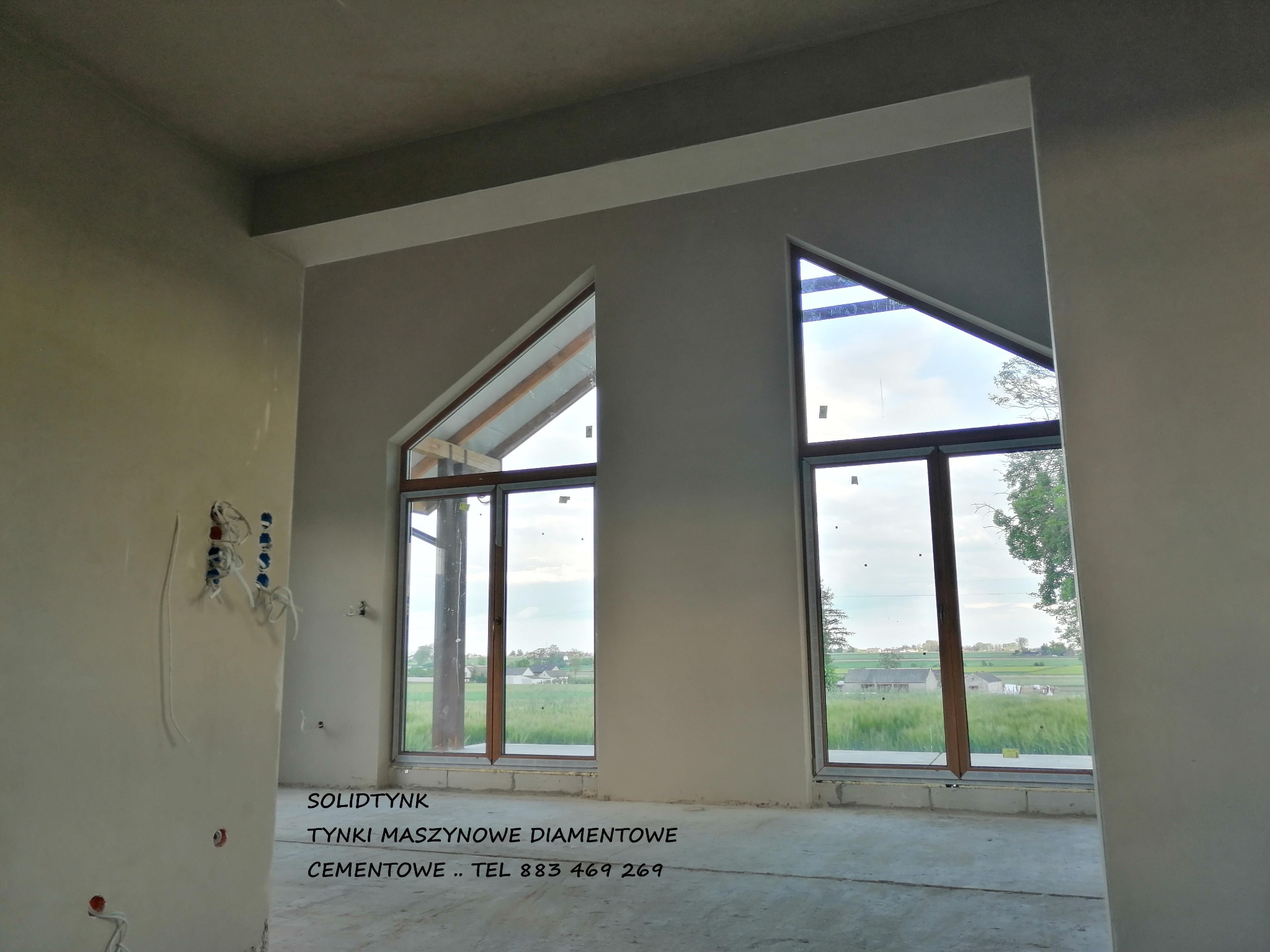 Wnętrze domu po tynkowaniu maszynowym. Widoczne okna z trójkątnymi nadprożami i gładkie, jasne ściany. Instalacja elektryczna wystaje ze ściany.