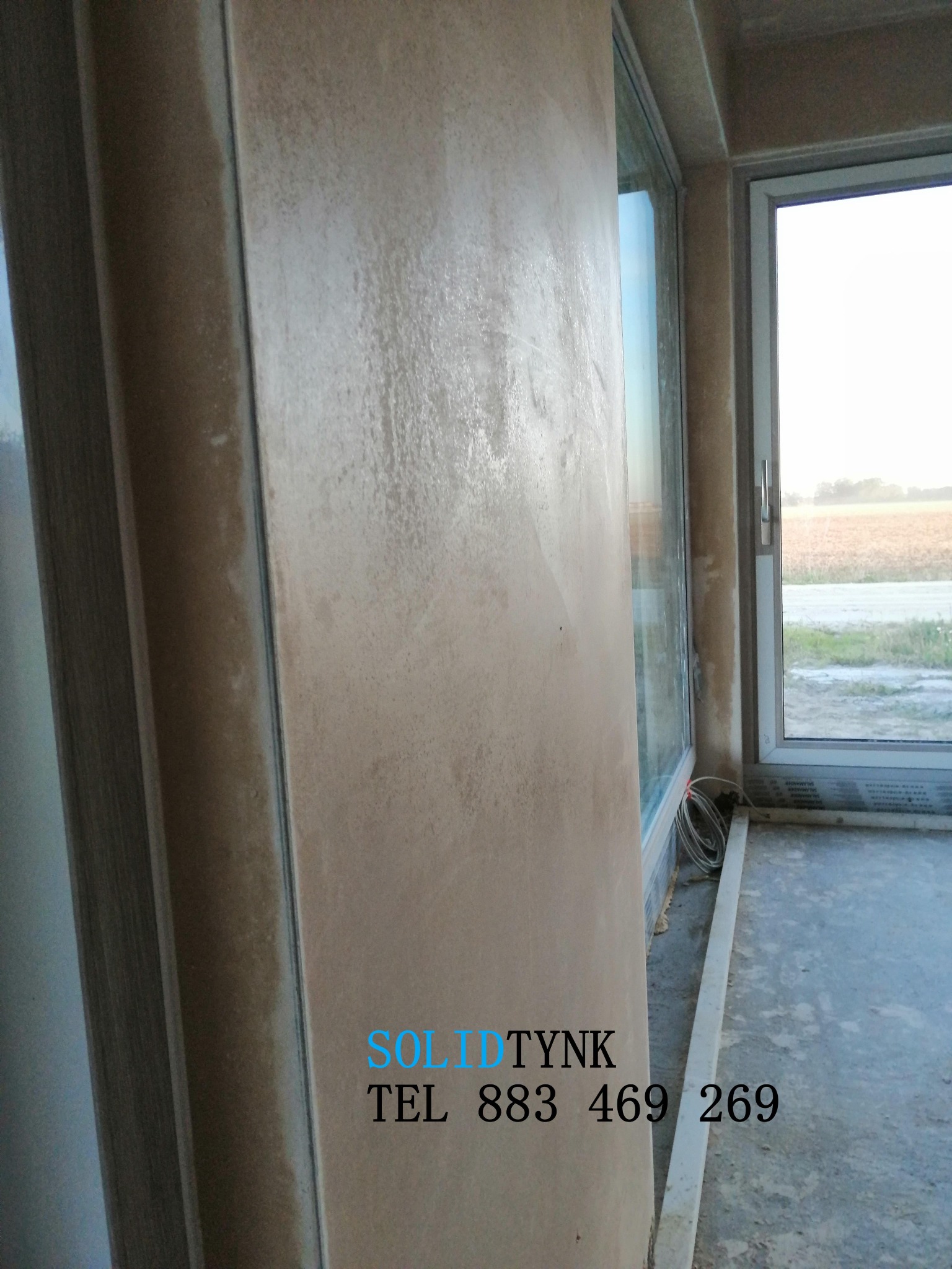 Ściana z efektem dekoracyjnym w surowym wnętrzu, widok z boku, z oknem w tle. Widoczne przewody i listwa przypodłogowa. Na ścianie logo firmy.