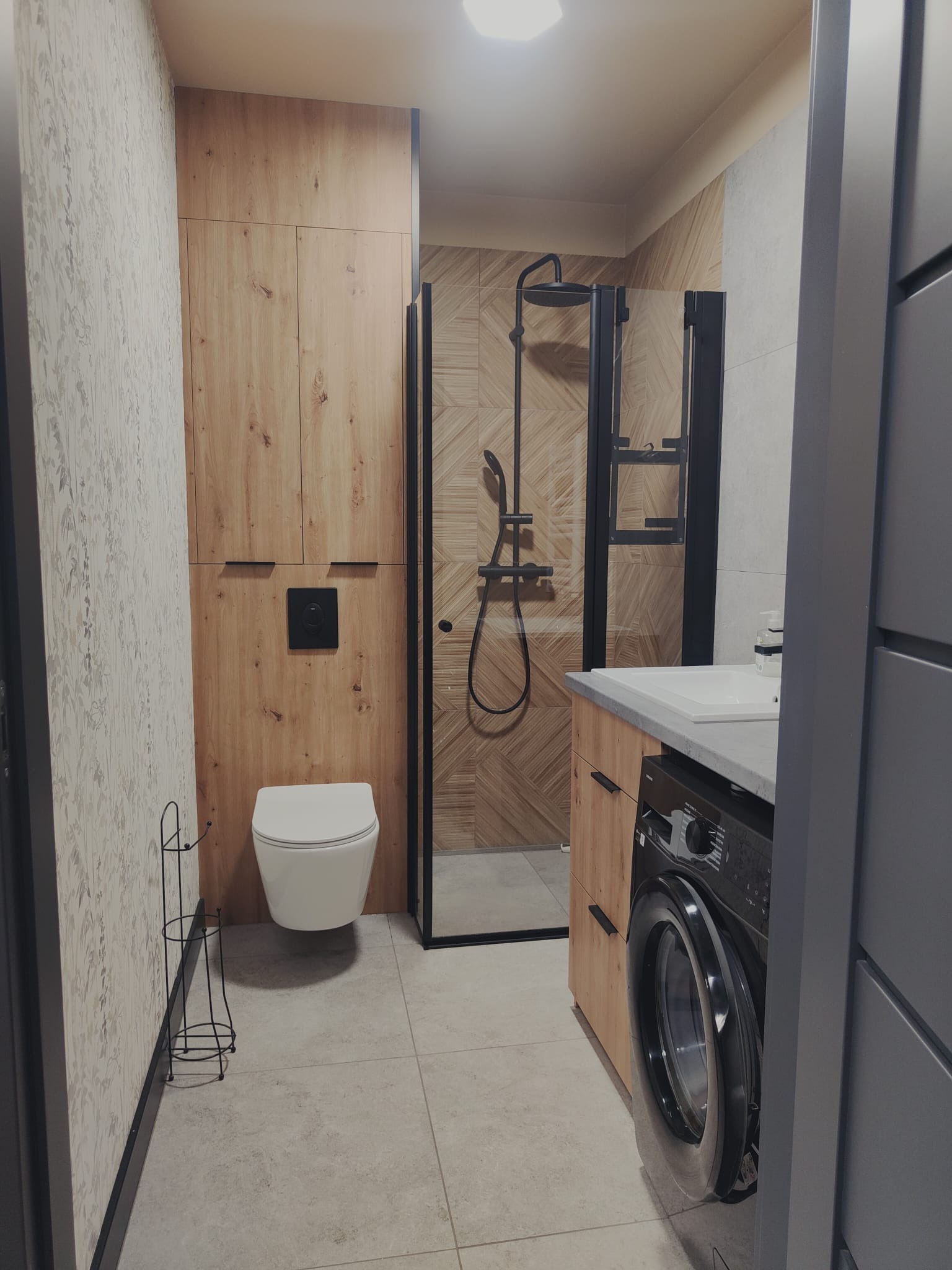 Nowoczesna łazienka z kabiną prysznicową typu walk-in, toaletą podwieszaną i pralką pod blatem umywalki. Drewniane szafki i geometryczne płytki dodają elegancji.