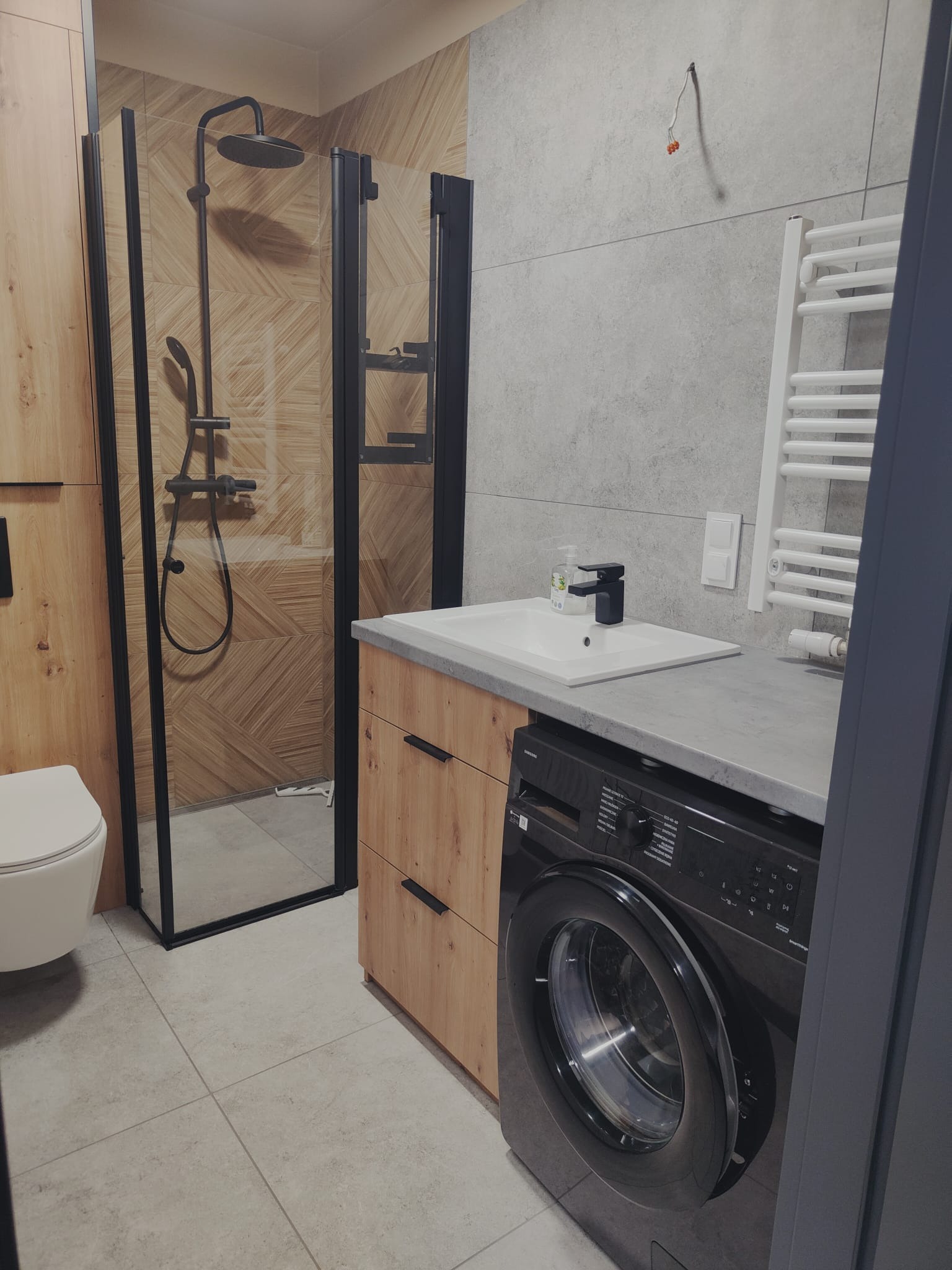 Nowoczesna łazienka z kabiną prysznicową typu walk-in, drewnianymi szafkami, szarą umywalką i czarną pralką. Minimalistyczny design, płytki imitujące beton.